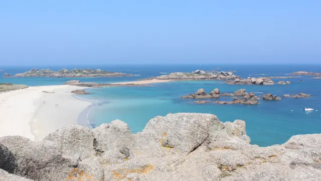 Blick auf einen weißen Sandstrand, der von türkisblauem Wasser und Felsen gesäumt wird, mit Segelbooten in der Ferne