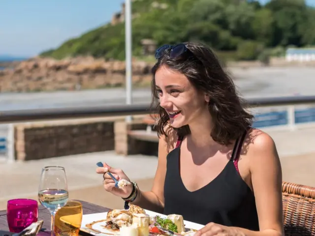 Le long d'un plage, une femme souriante assise en terrasse d'un restaurant, dégustant un plat de poisson.