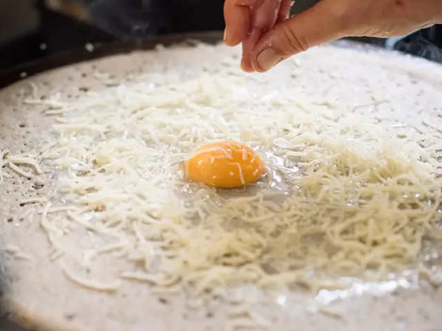 Une main dépose du fromage râpé sur une galette de blé noir en préparation sur une billig.