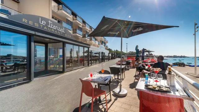 Terrasse extérieure d’un restaurant en bord de port avec tables, chaises et clients, sous des parasols