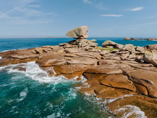 Sur la Côte de Granit Rose, un rocher en équilibre domine des rochers érodés par la mer, avec des vagues s’écrasant contre ce chaos granitique.