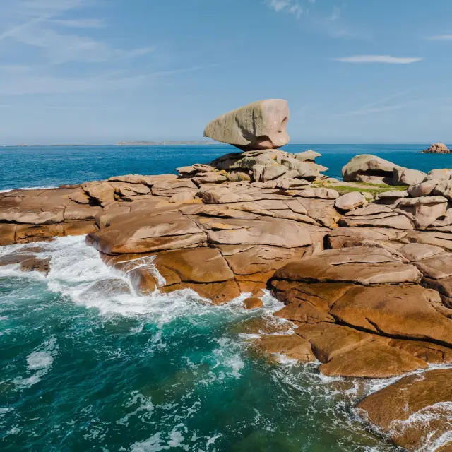 Sur la Côte de Granit Rose, un rocher en équilibre domine des rochers érodés par la mer, avec des vagues s’écrasant contre ce chaos granitique.
