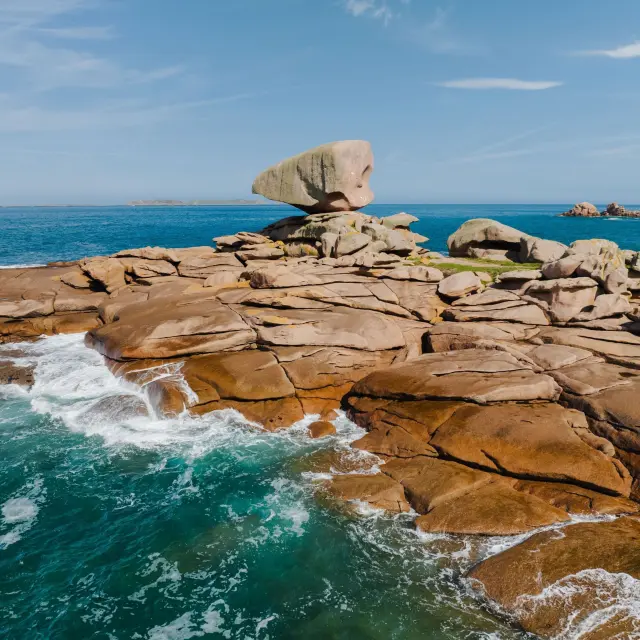 Sur la Côte de Granit Rose, un rocher en équilibre domine des rochers érodés par la mer, avec des vagues s’écrasant contre ce chaos granitique.