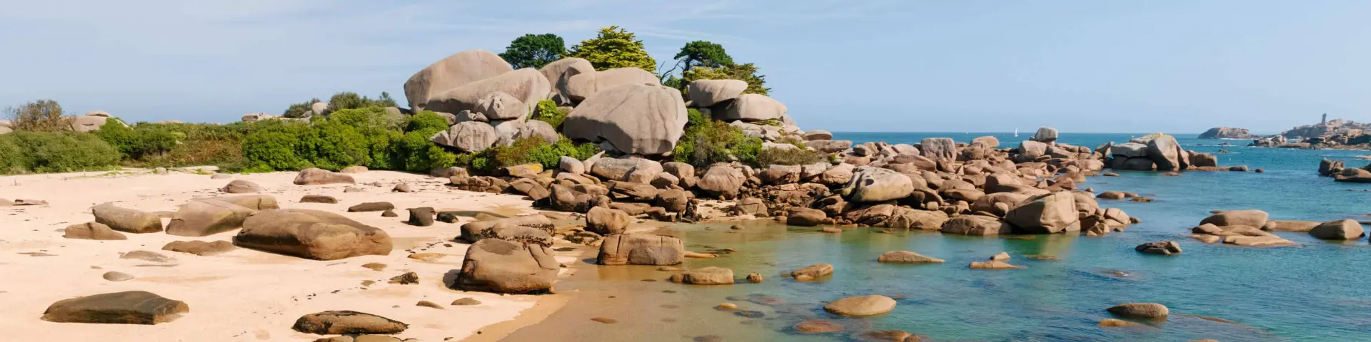 Un homme assis sur un chaos granitique contemple le paysage, sur une plage de sable rose bordée de rochers de granit rose aux formes arrondies, avec des reflets turquoise sur l’eau peu profonde.