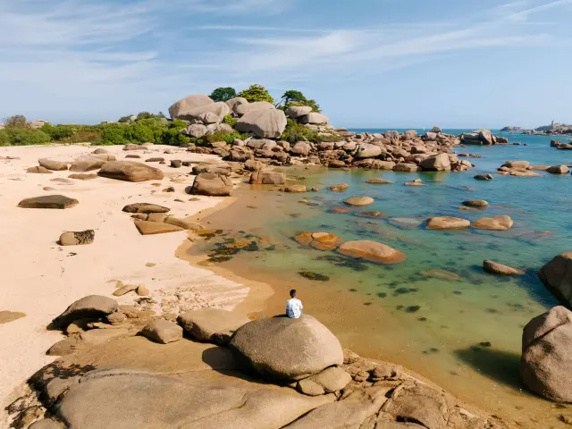 Weißer Sandstrand mit abgerundeten Felsen, türkisfarbene Reflexionen im flachen Wasser