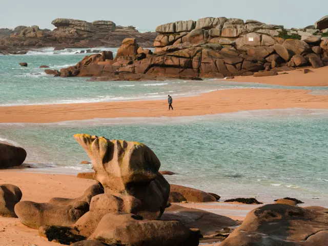 Sandy beach with rocks in the background and moored sailboats in turquoise water at sunset
