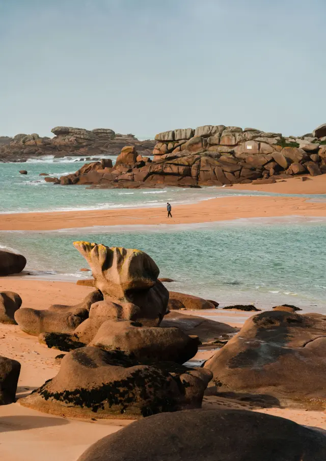 Sandy beach with rocks in the background and moored sailboats in turquoise water at sunset