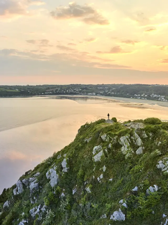 Vue panoramique du haut d'un grand rocher habillé de verdure sur une plage qui reflètent la lumière douce orangée du crépuscule.