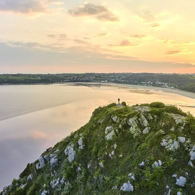 Vue panoramique du haut d'un grand rocher habillé de verdure sur une plage qui reflètent la lumière douce orangée du crépuscule.